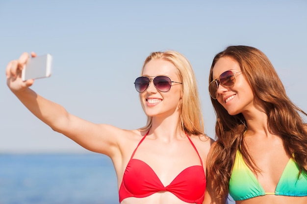 letnie wakacje, wakacje, podróże, technologia i koncepcja ludzi - dwie uśmiechnięte młode kobiety na plaży robiące selfie ze smartfonem na tle błękitnego nieba