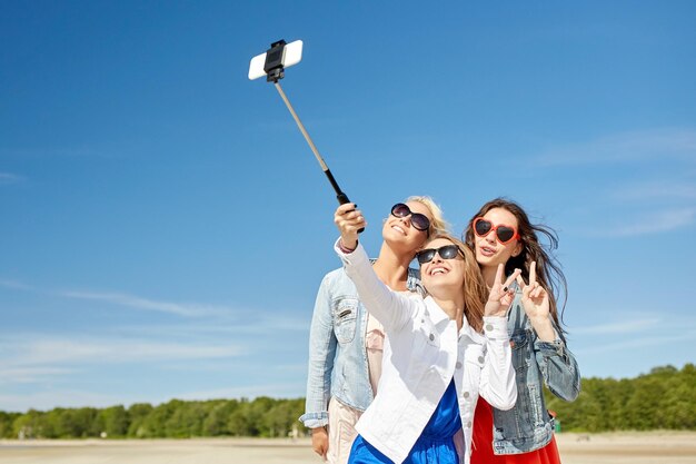 letnie wakacje, wakacje, podróże, technologia i koncepcja ludzi - grupa uśmiechniętych młodych kobiet robiących zdjęcia smartfonem na kiju do selfie na plaży