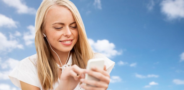 letnie wakacje, wakacje, technologia i koncepcja ludzi - uśmiechnięta młoda kobieta w białej sukni ze smartfonem i słuchawkami słuchająca muzyki na tle błękitnego nieba i chmur
