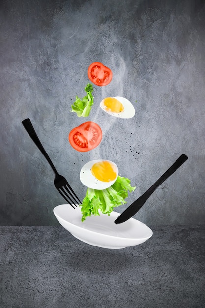 Lewitująca jajecznica z parą, pokrojonymi pomidorami, sałatą, białym talerzem, czarnym widelcem i nożem na szarym tle