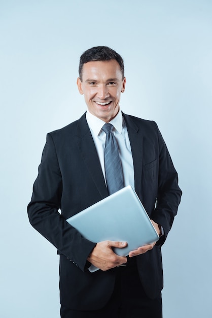 Lider biznesowy. Wesoły, zachwycony pozytywny człowiek uśmiecha się i trzyma laptopa, stojąc na niebieskim tle