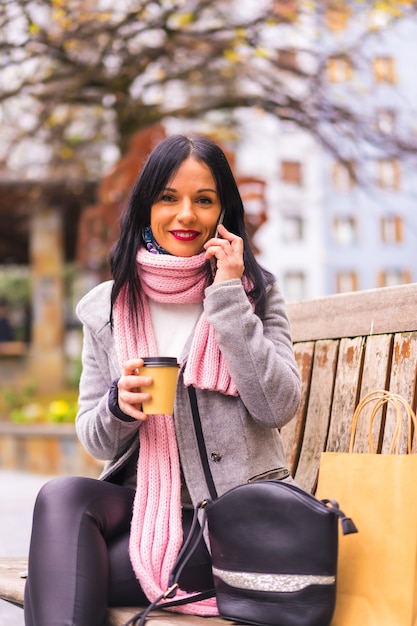 Lifestyle, kaukaski brunetka dziewczyna robi zakupy w mieście z papierowymi torbami i kawą na wynos, uśmiechnięta siedząca na ławce