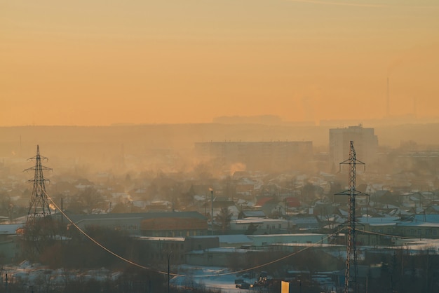 Linie energetyczne w mieście o świcie. Sylwetki budynków miejskich wśród smogu na wschód słońca. Kable wysokiego napięcia na ciepłym pomarańczowo-żółtym niebie.