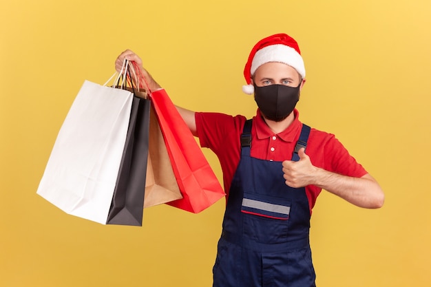 Listonosz w kapeluszu Świętego Mikołaja pokazujący kciuki w górę, trzymający torby na zakupy, kwarantanna dostawy towarów