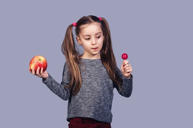 Little cute girl z cukierkiem i jabłkiem w dłoniach. Wybór koncepcji między zdrowym a niezdrowym jedzeniem. odizolowane na szarej powierzchni