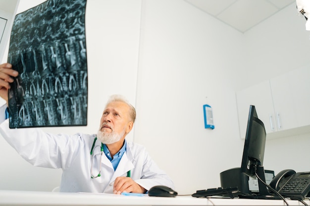 Lowangle widok poważnego starszego dorosłego lekarza płci męskiej badającego obraz kręgosłupa rezonansu magnetycznego pacjenta siedzącego w
