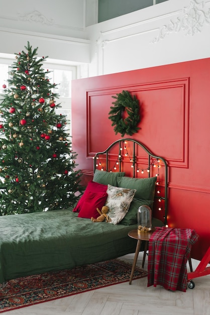 Łóżko z zielonymi poduszkami i prześcieradłem we wnętrzu świątecznej sypialni na tle czerwonej ściany z wieńcem Przy oknie stoi choinka