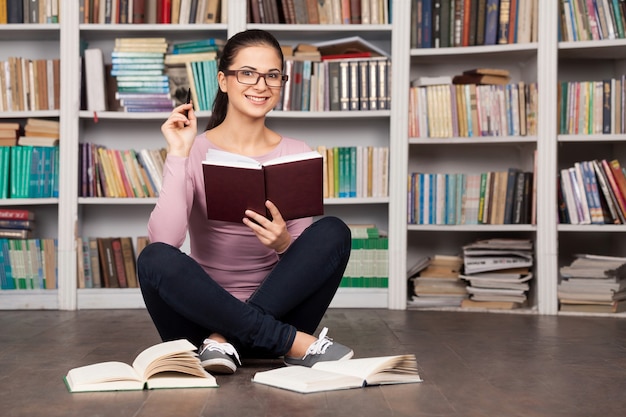 Lubi się uczyć. Wesoła młoda studentka trzymająca książki i uśmiechnięta siedząc na podłodze w bibliotece