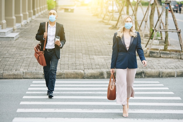 Ludzie biznesu noszący maski ochronne podczas chodzenia po ulicy i przechodzenia przez jezdnię