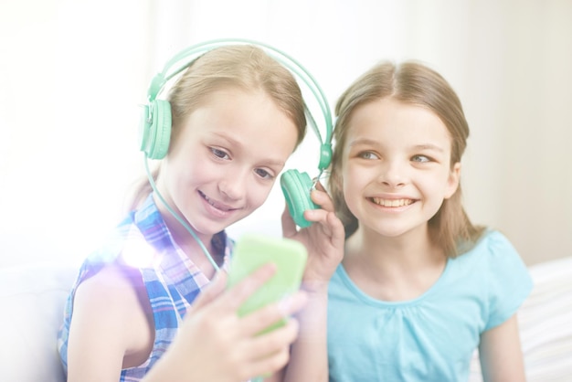 ludzie, dzieci, technologia, przyjaciele i koncepcja przyjaźni - szczęśliwe małe dziewczynki ze smartfonem i słuchawkami siedzą na kanapie i słuchają muzyki w domu