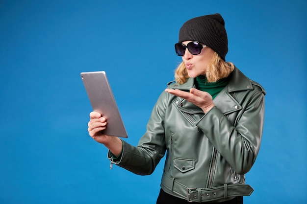 Ludzie gadżety i emocje koncepcja Całkiem urocza młoda dziewczyna w okularach przeciwsłonecznych i zieloną kurtkę wysyłając pocałunek powietrza podczas rozmowy wideo za pomocą cyfrowego tabletu na niebieskim tle studia
