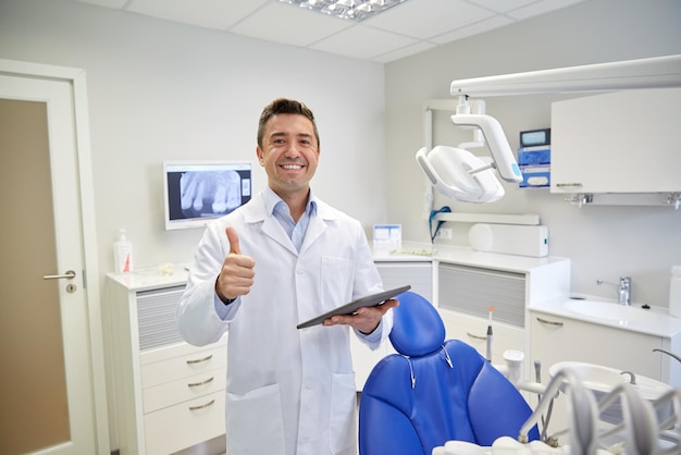 ludzie, medycyna, stomatologia i koncepcja opieki zdrowotnej - szczęśliwy dentysta mężczyzna w średnim wieku w białym fartuchu z komputerem typu tablet pc pokazujący kciuki do góry w biurze kliniki dentystycznej