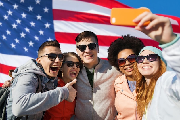 ludzie, międzynarodowa przyjaźń i koncepcja technologii - grupa szczęśliwych nastoletnich przyjaciół biorących selfie ze smartfonem i pokazujących kciuki w górę na tle flagi amerykańskiej