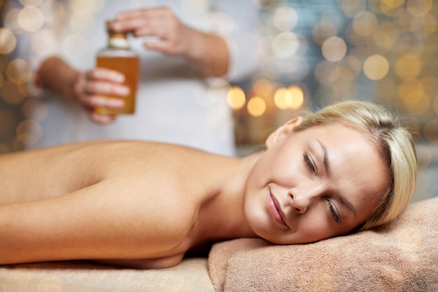 Zdjęcie ludzie, piękno, spa, zdrowy styl życia i koncepcja relaksu - zbliżenie pięknej młodej kobiety leżącej z zamkniętymi oczami na stole do masażu i terapeuty trzymającego butelkę oleju w spa