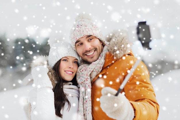 ludzie, pora roku, miłość, technologia i koncepcja wypoczynku - szczęśliwa para robi zdjęcie smartfonem na kiju selfie na zimowym tle