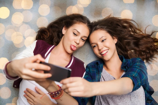 ludzie, przyjaciele, nastolatki i koncepcja przyjaźni - szczęśliwe uśmiechnięte ładne nastolatki leżące na podłodze i robiące selfie ze smartfonem na tle świateł świątecznych