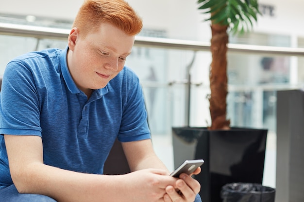 Ludzie, rozrywka i nowoczesna technologia. Młody czerwony kierowniczy uczeń ma odpoczynek w centrum handlowym trzyma jego smartphone używać wysokiej prędkości połączenie z internetem. Stylowy rudy facet w niebieskiej koszulce, piszący na maszynie