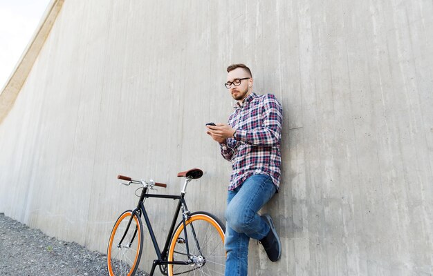 ludzie, styl, technologia, wypoczynek i styl życia - szczęśliwy młody hipster człowiek w słuchawkach ze smartfonem i rowerem stacjonarnym, słuchając muzyki na ulicy miasta