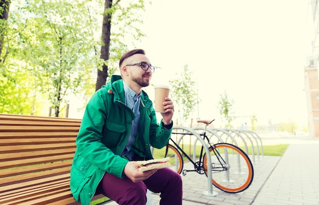 ludzie, styl, wypoczynek i styl życia - szczęśliwy młody hipster z kanapką, filiżanką kawy i rowerem z ostrym kołem zaparkowanym na ulicy miasta
