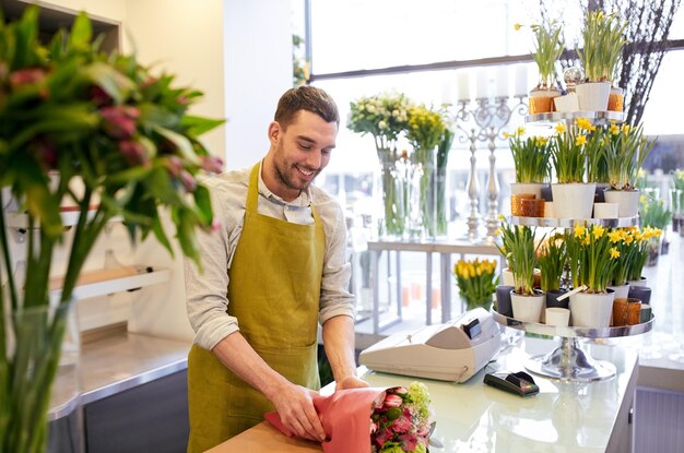 ludzie, zakupy, sprzedaż, florystyka i koncepcja konsumpcjonizmu - szczęśliwy uśmiechnięty kwiaciarz zawijający kwiaty w papier w kwiaciarni
