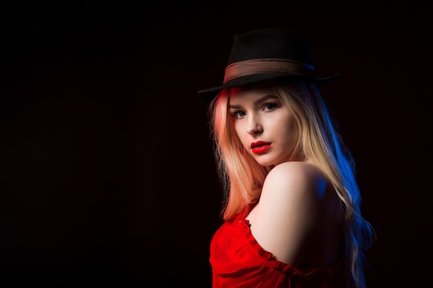 Luksusowa blondynka w czerwonej bluzce i brązowym kapeluszu pozuje w studio z cieniem na twarzy