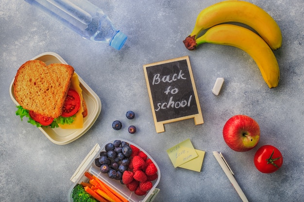 Lunchbox z kanapek jagod marchewek brokułów butelką wodny banan na szarym odgórnym widoku