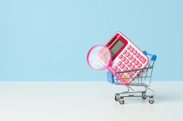 Lupa i różowy plastikowy kalkulator w miniaturowym różowym wózku na niebieskim tle. Koncepcja planowania budżetu, liczenie oszczędności, kontrola kosztów
