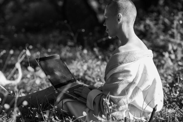 Łysa kobieta w szlafroku siedzi na zielonej trawie z laptopem w dłoniach Koncepcja niezależnej pracy online Słoneczny dzieńCzarno-biały obraz