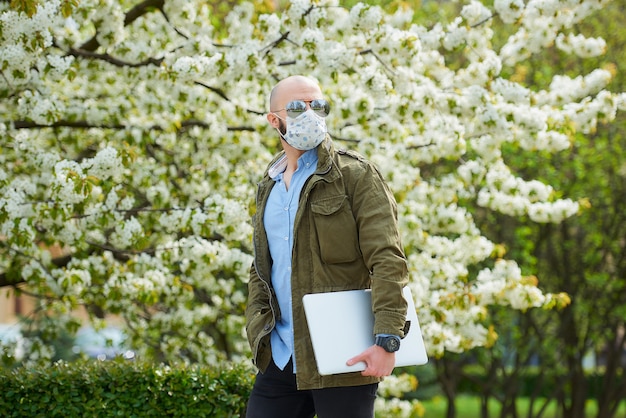 Łysy mężczyzna z brodą w medycznej masce na twarz, aby uniknąć rozprzestrzeniania się koronawirusa, idzie z laptopem w parku. Facet nosi maskę N95 i pilotowe okulary przeciwsłoneczne w ogrodzie między kwitnącymi drzewami.