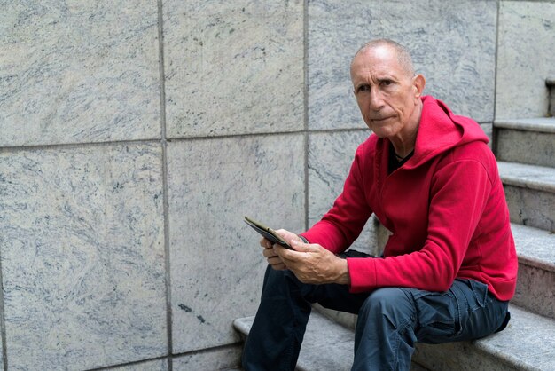 Łysy starszy mężczyzna myśli i trzymając cyfrowy tablet, siedząc na schodach