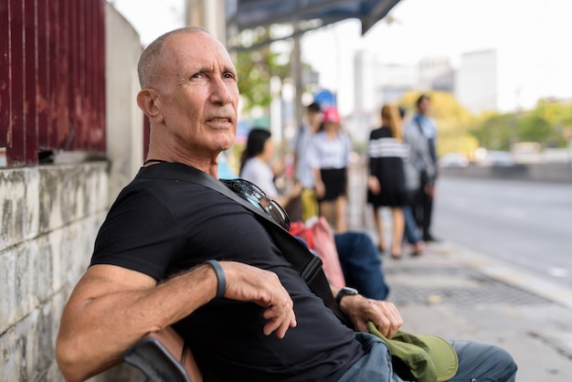 Łysy starszy turysta czeka i siedzi na drewnianej ławce na przystanku autobusowym