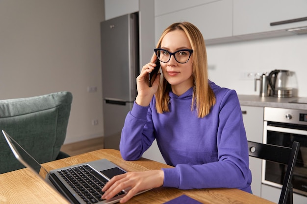 M?oda doros?a kobieta w fioletowej bluzie z kapturem rozmawia przez telefon komórkowy i korzysta z laptopa Koncepcja pracy w domu