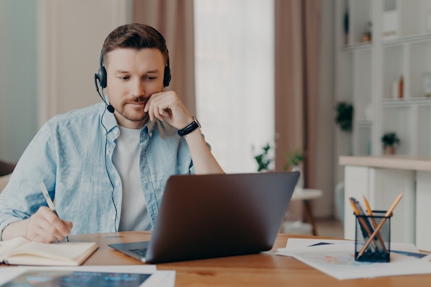 Mądry freelancer słucha ważnych informacji online, robi niezbędne notatki w pamiętniku, pracuje na laptopie w domu, na komputerze, nosi dżinsową koszulę na rozmytym tle. Usługa wsparcia przez infolinię