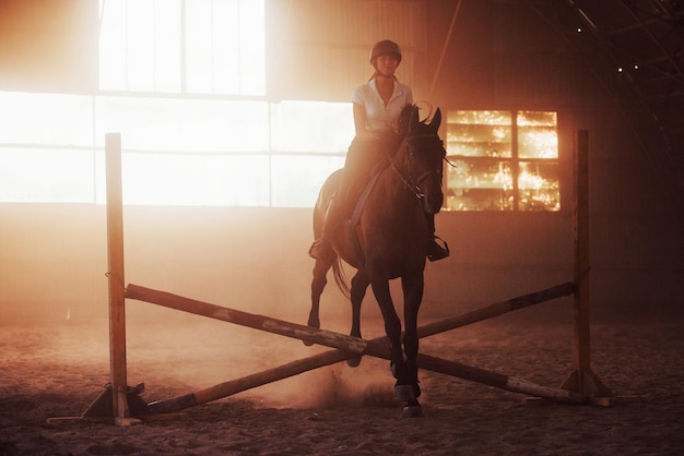 Majestatyczny obraz sylwetki konia z jeźdźcem na tle zachodu słońca. Dżokejka na grzbiecie ogiera jeździ w hangarze na farmie.
