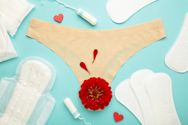 Majtki damskie z wkładkami menstruacyjnymi i tamponami na pastelowym niebieskim tle. Widok z góry. Pojęcie dni krytycznych, miesiączki, higieny kobiecej. Kwiat róży leży na poduszce menstruacyjnej.