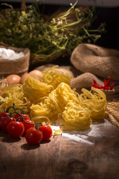 Makaron fettuccine na drewnianym stole z pomidorkami koktajlowymi, papryką, mąką pszenną i jajkami w tle.