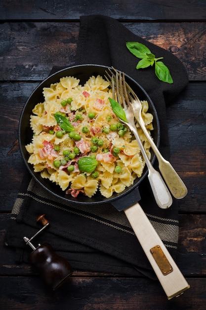 Makaron fettuccine z boczkiem i zielonym groszkiem i parmezanem na starej patelni na starej ciemnej powierzchni drewnianej