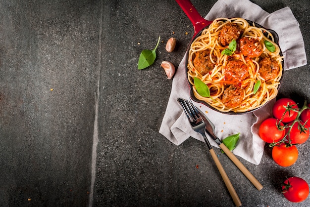 Makaron spaghetti z klopsikami, bazyliowym sosem pomidorowym w czerwonej żeliwnej patelni, na czarnym kamiennym stole