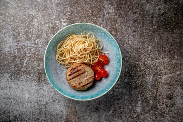 Makaron spaghetti z pasztecikiem mięsnym i świeżymi pomidorami na niebieskim talerzu na tle szarego kamiennego stołu.