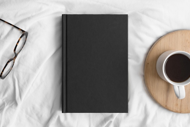 Makieta czarnej książki z filiżanką kawy i okularami na łóżku