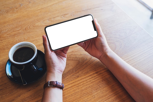 Makieta obraz kobiety trzymającej czarny telefon komórkowy z pustym ekranem z filiżanką kawy na drewnianym stole