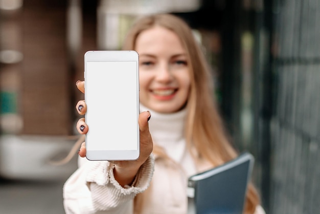 Makieta obrazu blond studentki pokazującej telefon komórkowy z pustym białym ekranem na tle budynku uniwersyteckiego