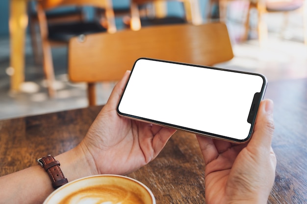 Makieta obrazu kobiety trzymającej czarny telefon komórkowy z pustym ekranem z filiżanką kawy na drewnianym stole