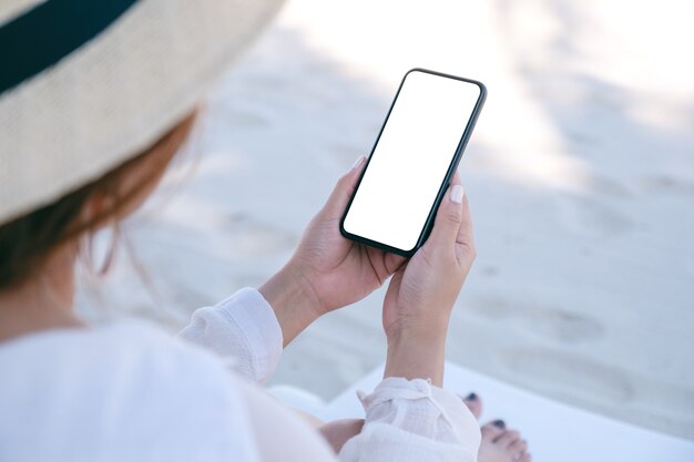 Makieta obrazu kobiety trzymającej i używającej białego telefonu komórkowego z pustym ekranem pulpitu siedząc na leżaku na plaży