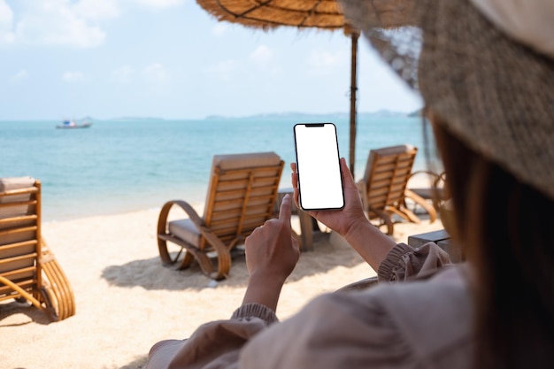 Makieta obrazu kobiety trzymającej telefon komórkowy z pustym ekranem pulpitu podczas siedzenia na plaży