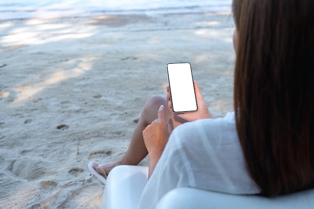 Makieta obrazu kobiety trzymającej telefon komórkowy z pustym ekranem pulpitu podczas siedzenia na plaży