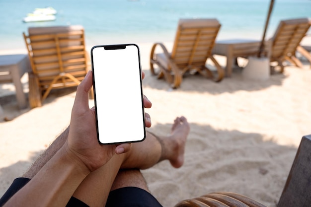 Makieta obrazu mężczyzny trzymającego telefon komórkowy z pustym ekranem pulpitu podczas siedzenia na plaży