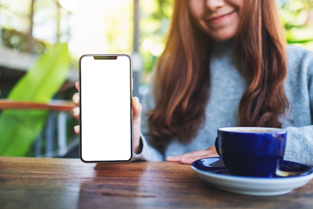 Makieta obrazu pięknej kobiety trzymającej i pokazującej czarny telefon komórkowy z pustym ekranem w kawiarni