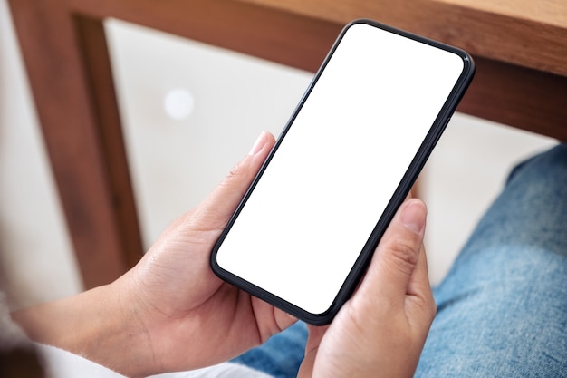 Makieta obrazu rąk trzymających czarny telefon komórkowy z pustym białym ekranem podczas siedzenia