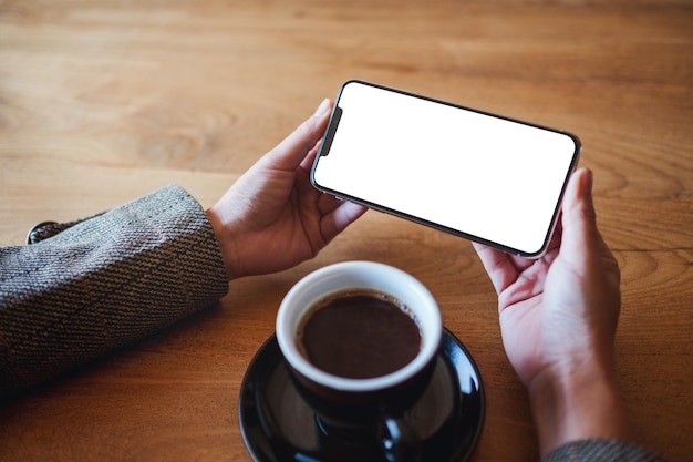 Makieta obrazu rąk trzymających telefon komórkowy z pustym ekranem z filiżanką kawy na drewnianym stole